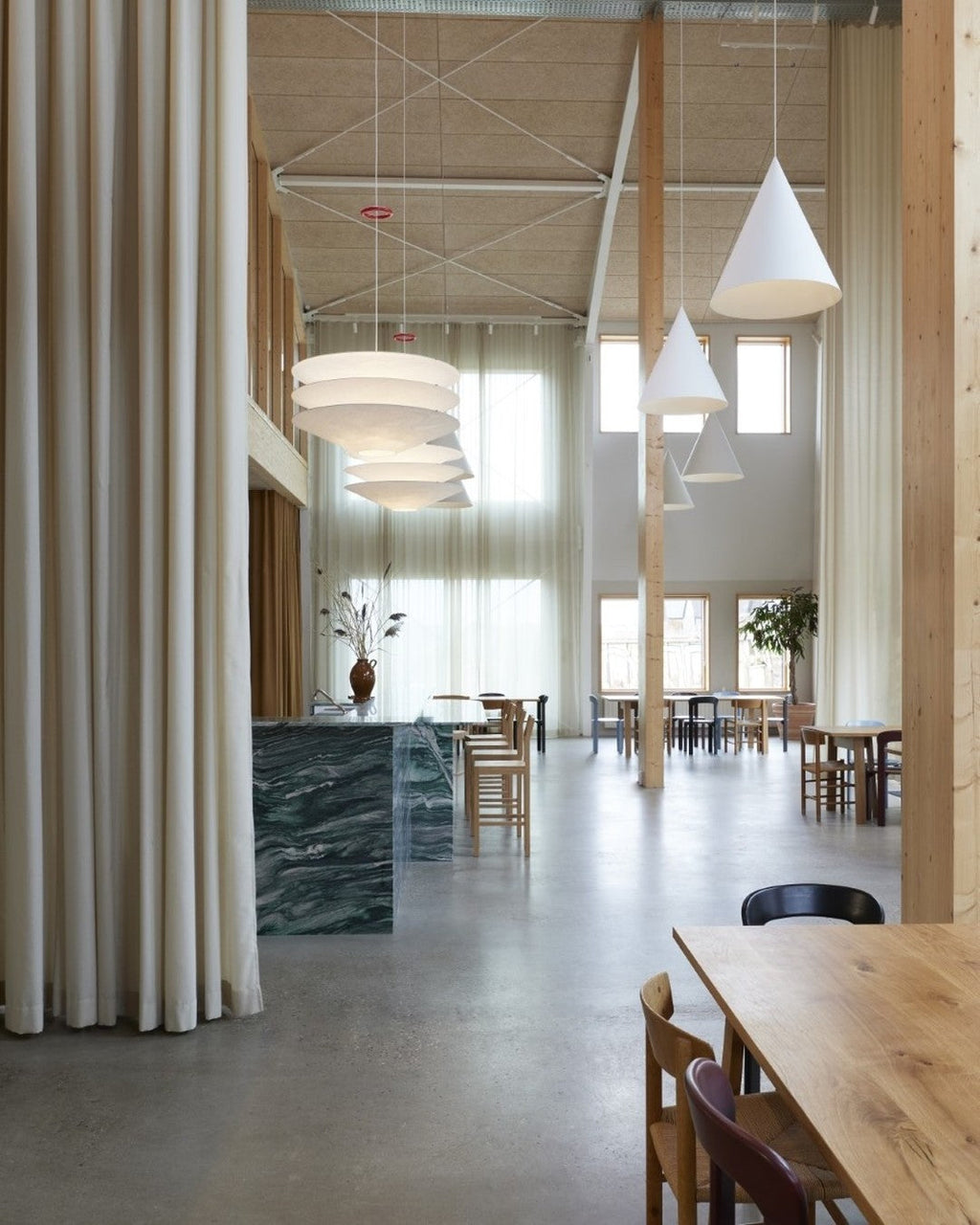 In a tall timber-framed room with floor-length curtains, tiered Clérachic pendants hang beside plain white cone pendants over a long table.