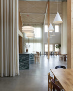 In a tall timber-framed room with floor-length curtains, tiered Clérachic pendants hang beside plain white cone pendants over a long table.