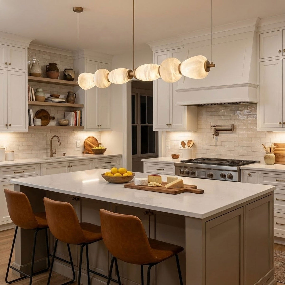 Cream kitchen with a Clérachic pebble chandelier running over an island with tan leather stools, lemons and a cheese board.
