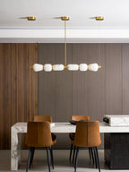 Higher view of this Clérachic linear chandelier with its three gold ceiling canopies, over a marble table and tan leather chairs.