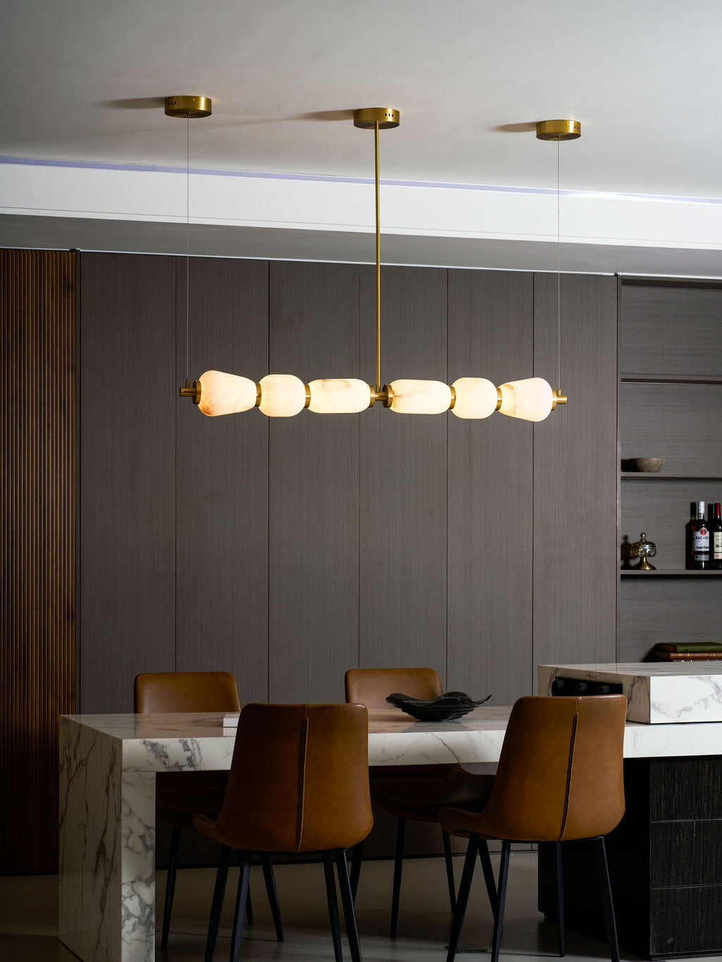 Evening view of a Clérachic linear chandelier lit warm amber above a marble table, with leather chairs and shelves of bottles behind.