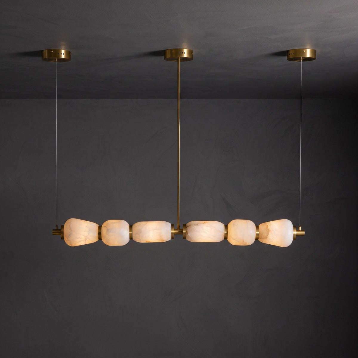 Lit six-pebble Clérachic chandelier with gold fittings hanging against a dark plaster wall, the alabaster glowing amber.