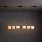 A Clérachic linear chandelier with black canopies, its six alabaster pebbles glowing warm against a dark textured wall.