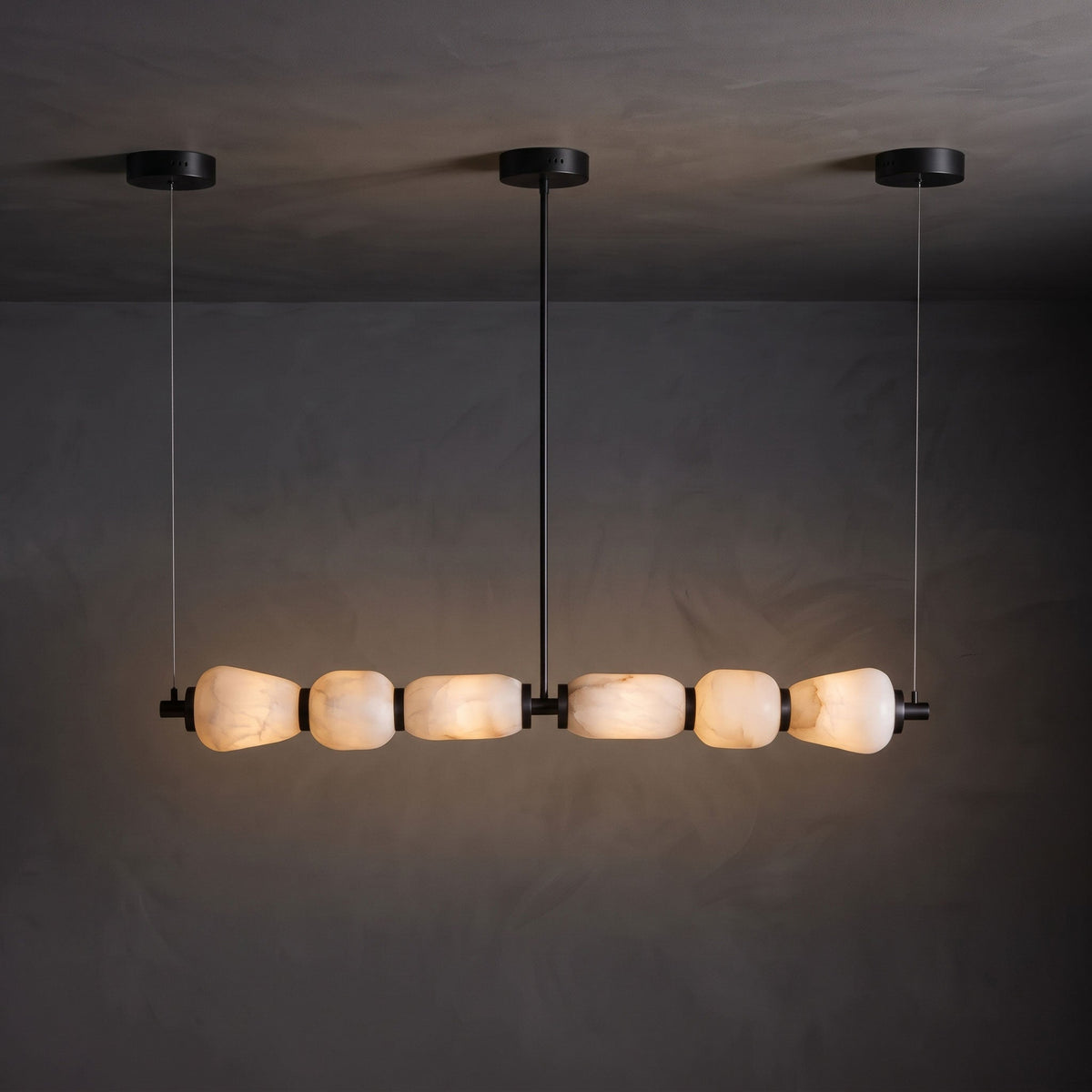 A Clérachic linear chandelier with black canopies, its six alabaster pebbles glowing warm against a dark textured wall.