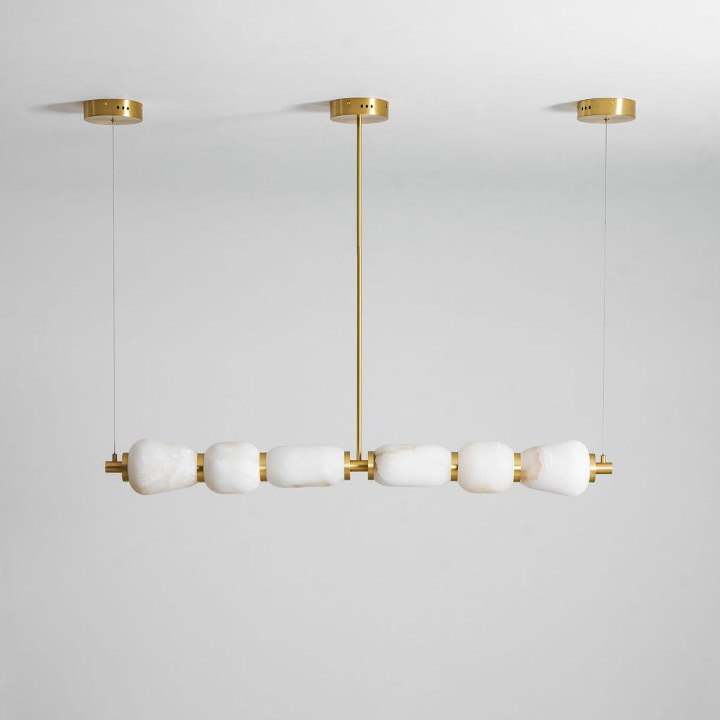 Unlit Clérachic linear chandelier in gold and white, with six alabaster pebbles under three gold canopies against a pale grey wall.