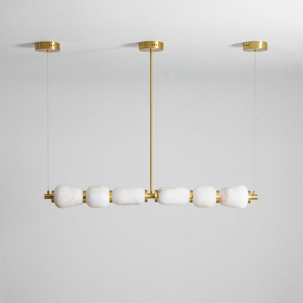 Unlit Clérachic linear chandelier in gold and white, with six alabaster pebbles under three gold canopies against a pale grey wall.
