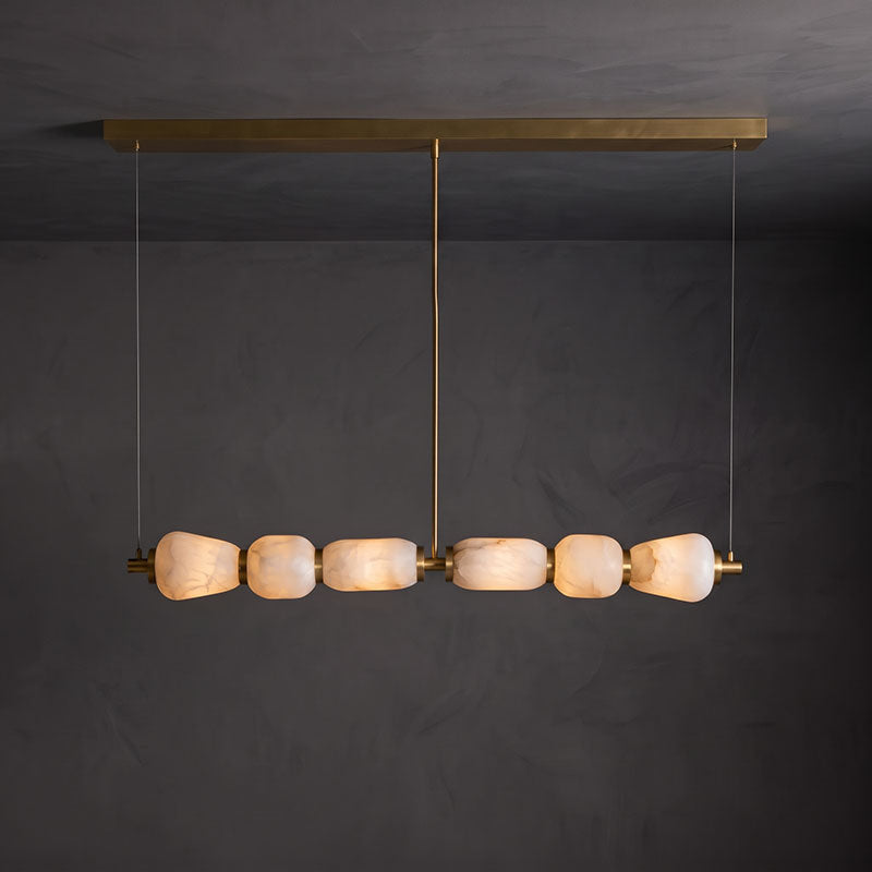 Clérachic linear chandelier with a long gold canopy bar, its six alabaster pebbles lit warm against a dark grey wall.