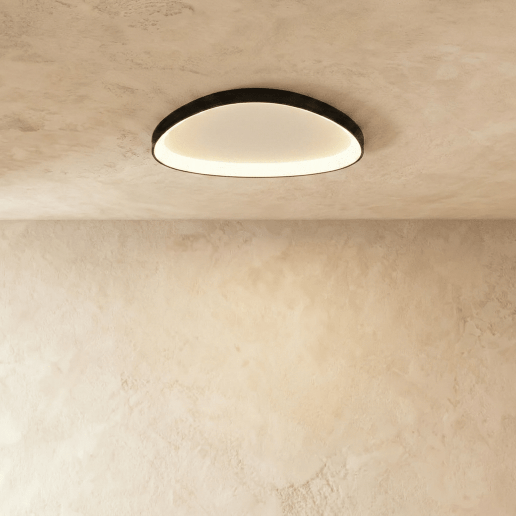 Clérachic black aluminium ceiling light, a soft rounded oval, glowing warm from its inner rim on a limewashed ceiling.