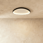 Clérachic black aluminium ceiling light, a soft rounded oval, glowing warm from its inner rim on a limewashed ceiling.