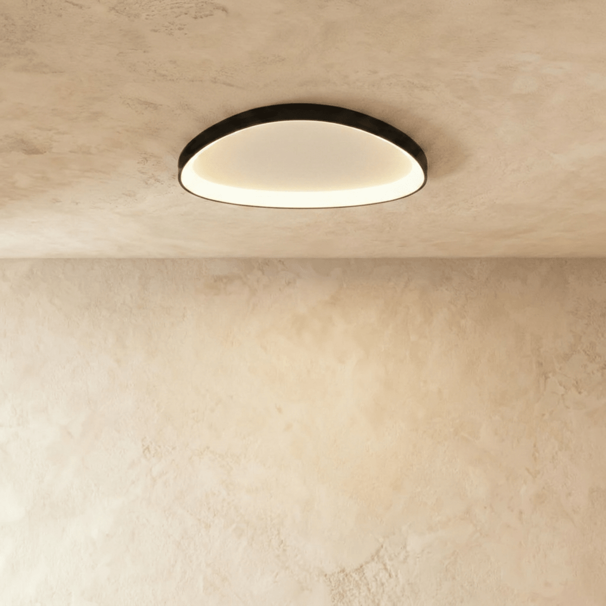 Clérachic black aluminium ceiling light, a soft rounded oval, glowing warm from its inner rim on a limewashed ceiling.