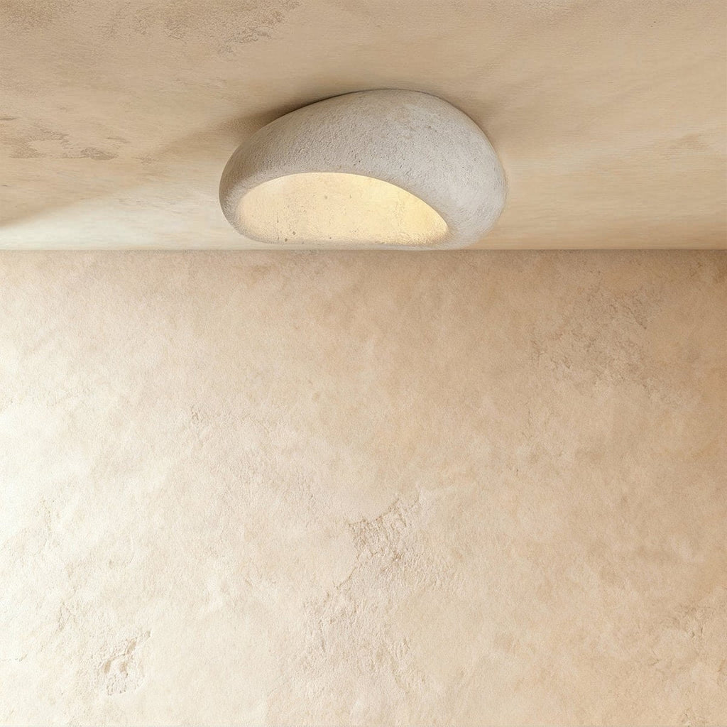 Clérachic pebble-shaped flush ceiling light in a pale speckled cement finish, with warm light glowing from its underside.