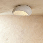 Clérachic mid-size pebble flush ceiling light in speckled cement white, lit warm and mounted on a plaster ceiling.