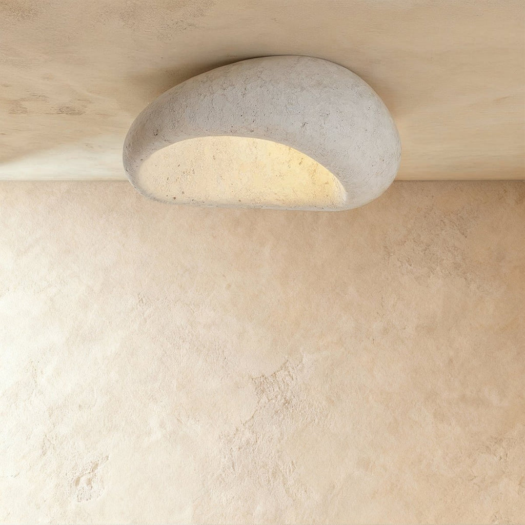 Clérachic larger pebble flush ceiling light in a pitted cement-white finish, its curved opening glowing warm.