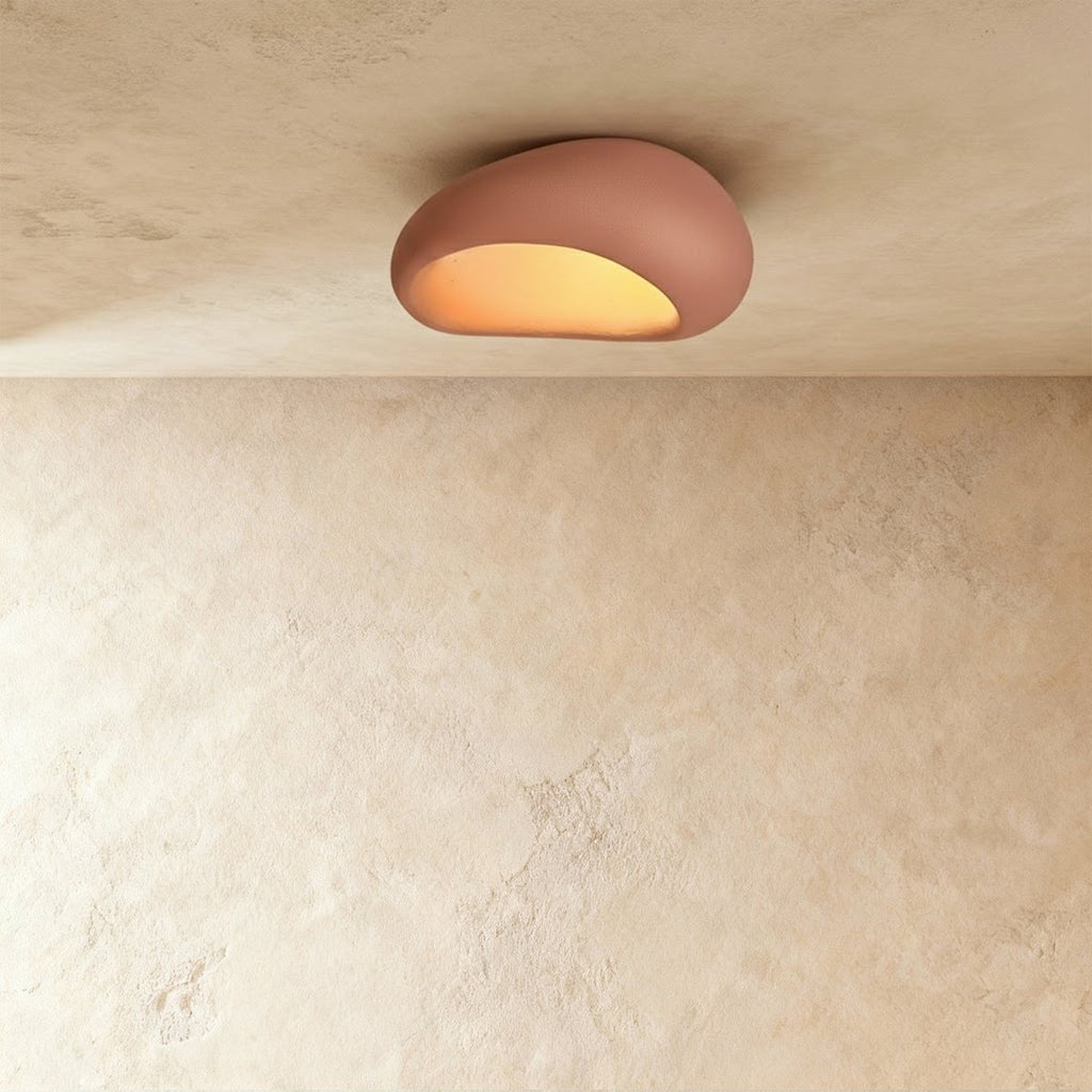 Clérachic small terracotta pink pebble flush ceiling light glowing amber from its curved opening on a plaster ceiling.