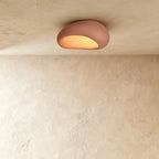 Clérachic small terracotta pink pebble flush ceiling light glowing amber from its curved opening on a plaster ceiling.