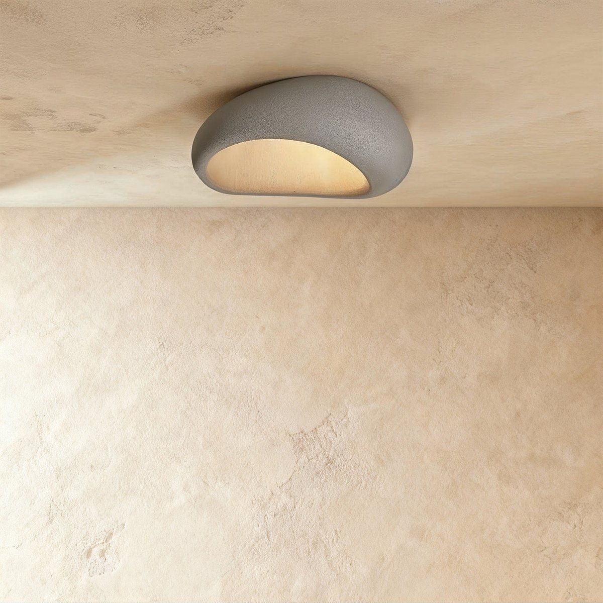 Clérachic mid-small grey pebble flush ceiling light with a speckled concrete-like shell, lit warm on a plaster ceiling.