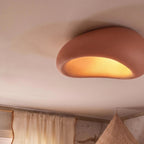 Close view of a Clérachic terracotta pink pebble ceiling light glowing amber in a nursery beside linen curtains.
