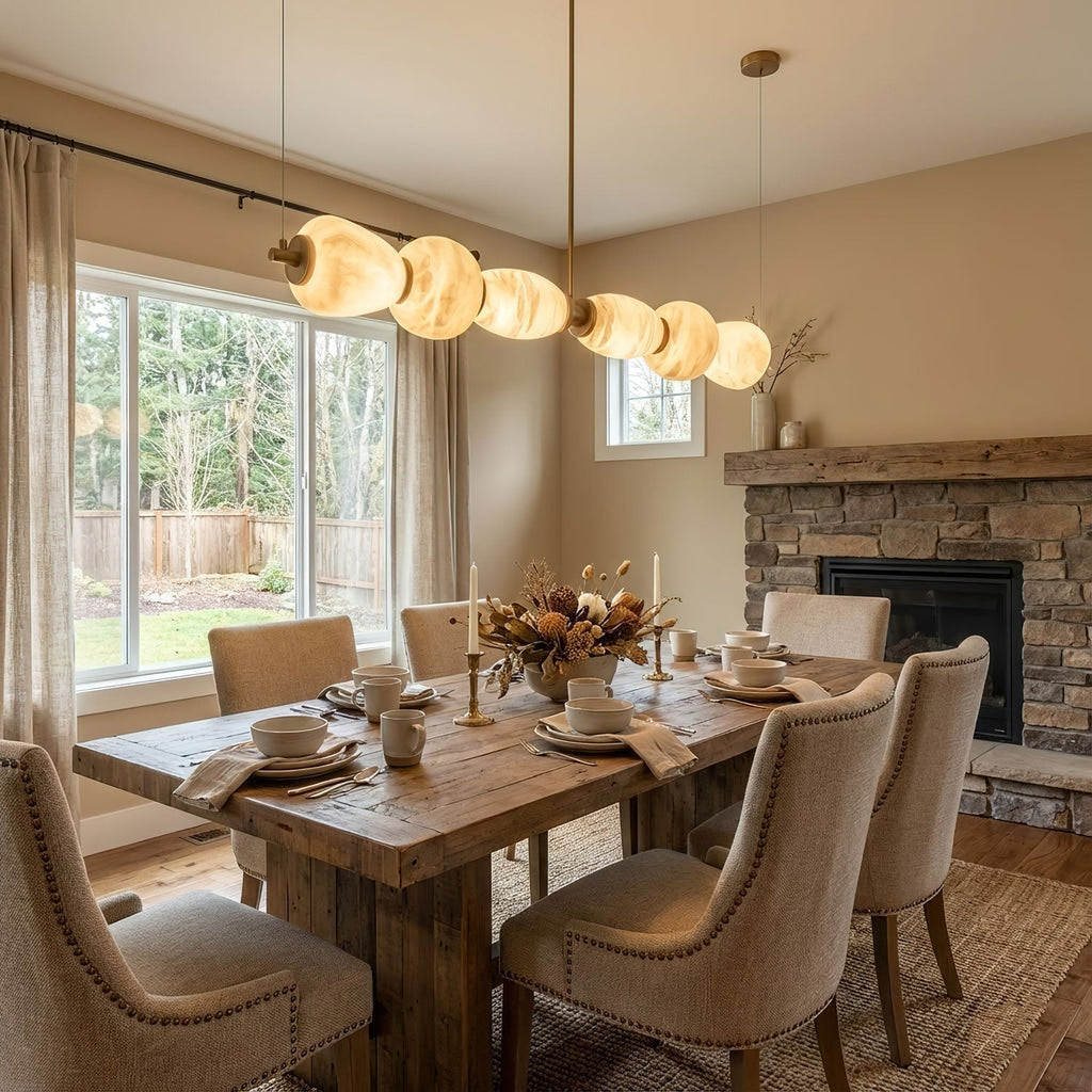 The Clérachic Pebble chandelier over a rustic dining table beside a stone fireplace and window.