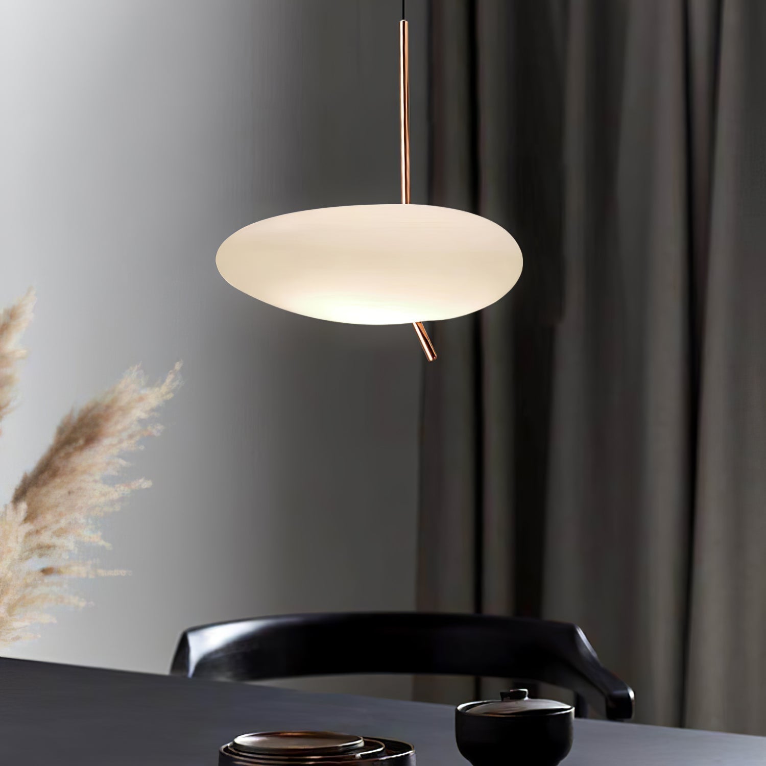 Clérachic pendant light glowing above a dark dining table set with small cups, framed by pampas grass and a curtain.