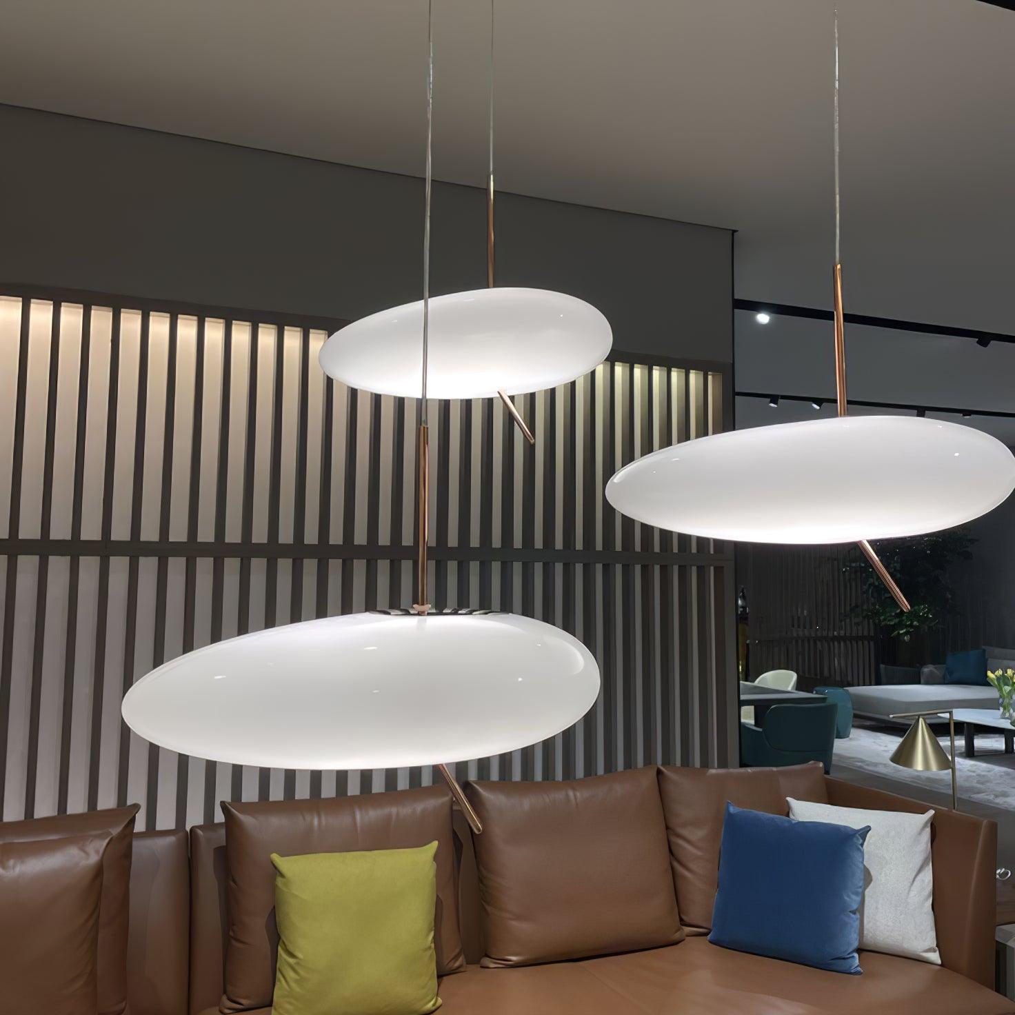 Three Clérachic pebble pendants glow above a brown leather sofa with colorful pillows and a slatted wood wall.