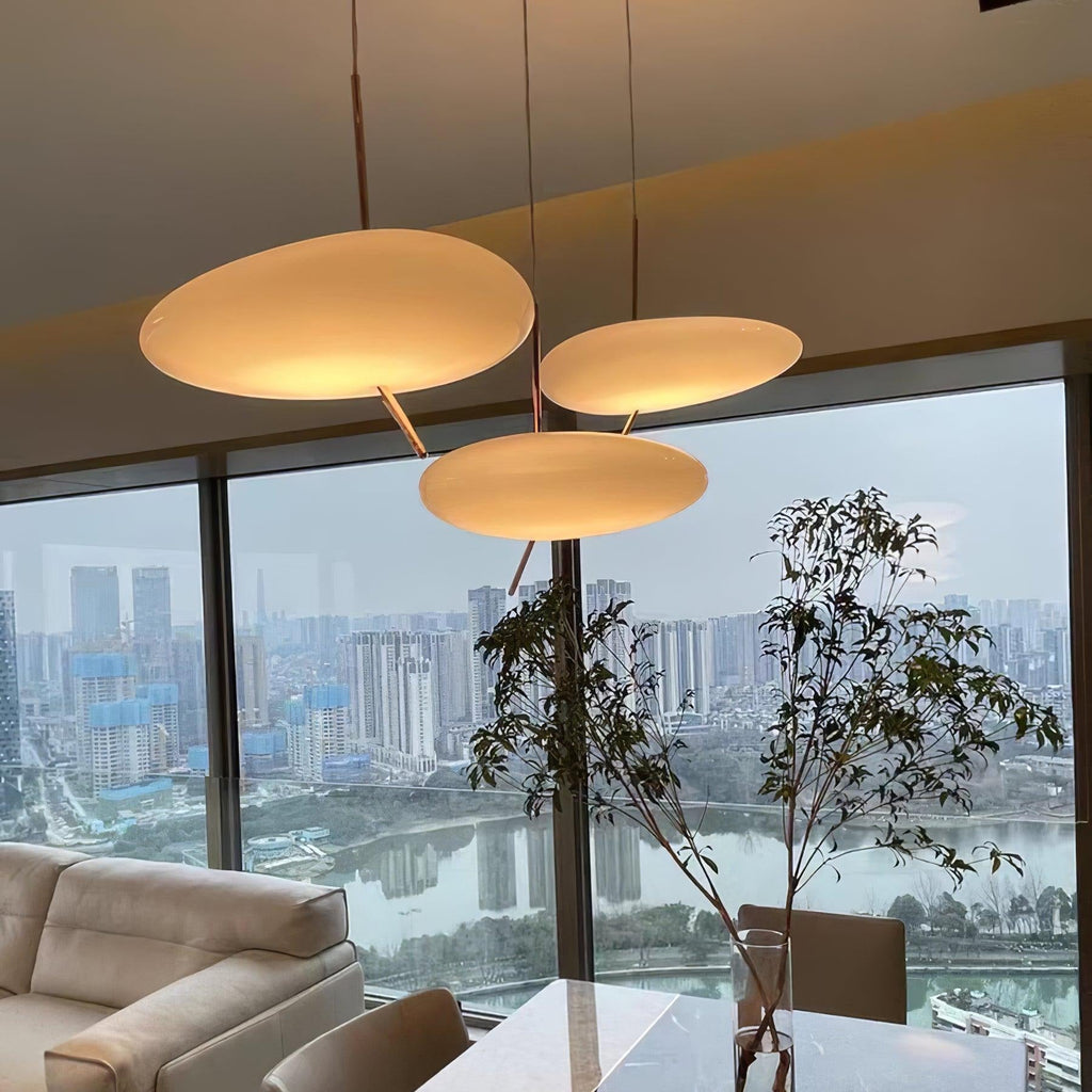 Three Clérachic pebble pendants glow over a dining table, framed by potted greenery and a city skyline view.
