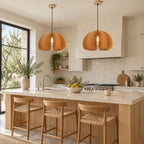 Two Clérachic slatted wood pendants lit over a pale kitchen island with woven-seat stools, a stone counter and a tall window.