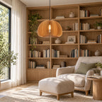 Clérachic wood slat pendant in front of a tall bookshelf, with an armchair, a pale ottoman and olive branches by the window.