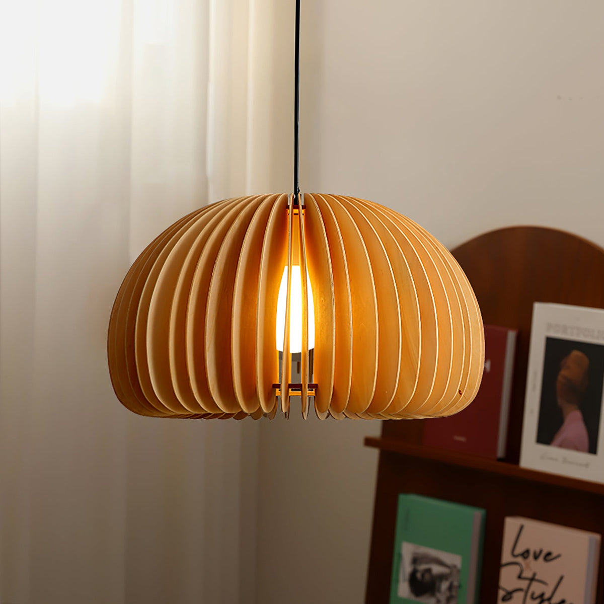 Close view of the vertical wood slats on this Clérachic pendant, with the bulb glowing inside, by a curtain and a shelf of books.
