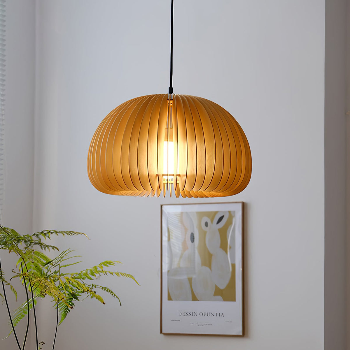 Lit Clérachic slatted pendant against a white wall, above a framed poster and a leafy plant, with the bulb visible through the slats.