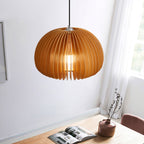 A Clérachic natural wood pendant hung in a corner by a window with a sheer curtain, above a wooden table with a magazine and a vase.