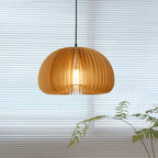 This Clérachic slatted pendant in front of a closed white blind, lit warmly, with fern fronds in the corner of the frame.
