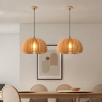 Two Clérachic wood slat pendants over a dining table with round-backed chairs, in front of a framed abstract print on a pale wall.