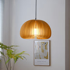 A Clérachic slatted pendant hung low in front of a pale wall, with a framed poster and green fronds behind it.