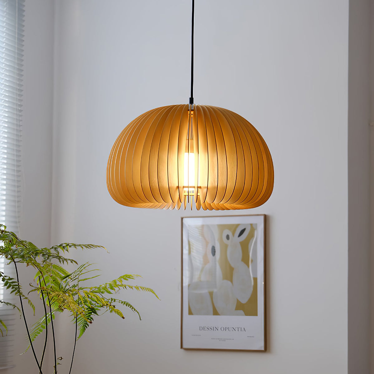 A Clérachic slatted pendant hung low in front of a pale wall, with a framed poster and green fronds behind it.
