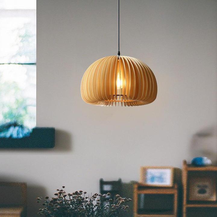 A Clérachic wood slat pendant lit in a living space, with a window and blurred shelves behind and a few small plants on a ledge.