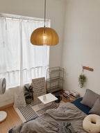 A Clérachic natural wood pendant in a bedroom with a white curtain, a low bed with grey bedding and a small clothes rail.