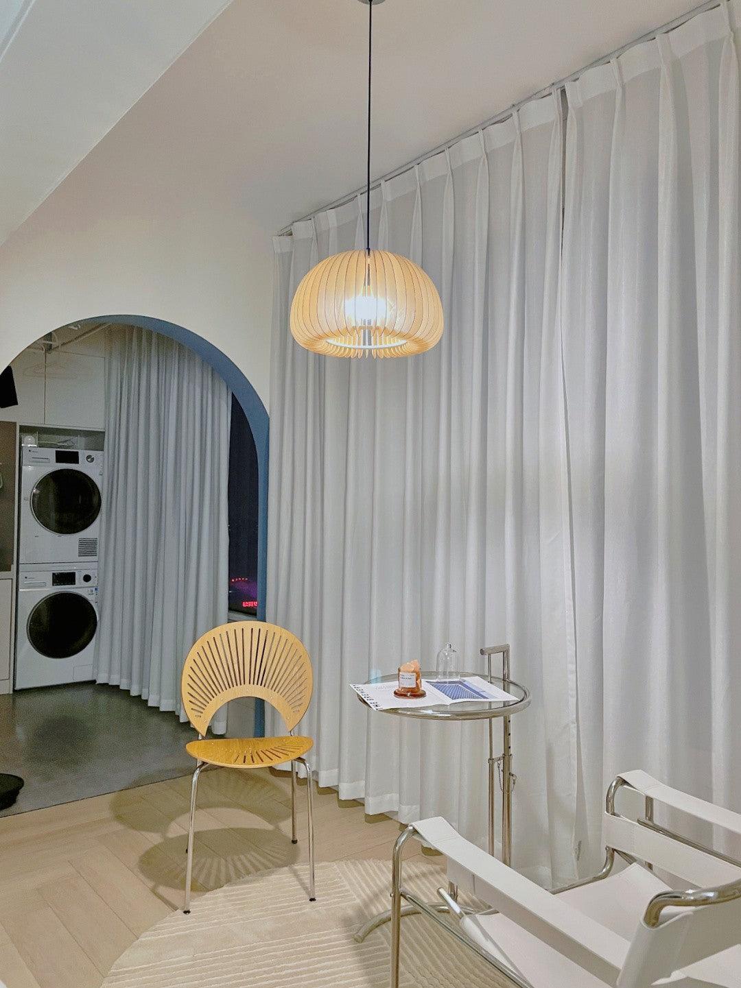 A Clérachic wood slat pendant hung in front of white curtains in an arched room, above a yellow shell-back chair and a side table.