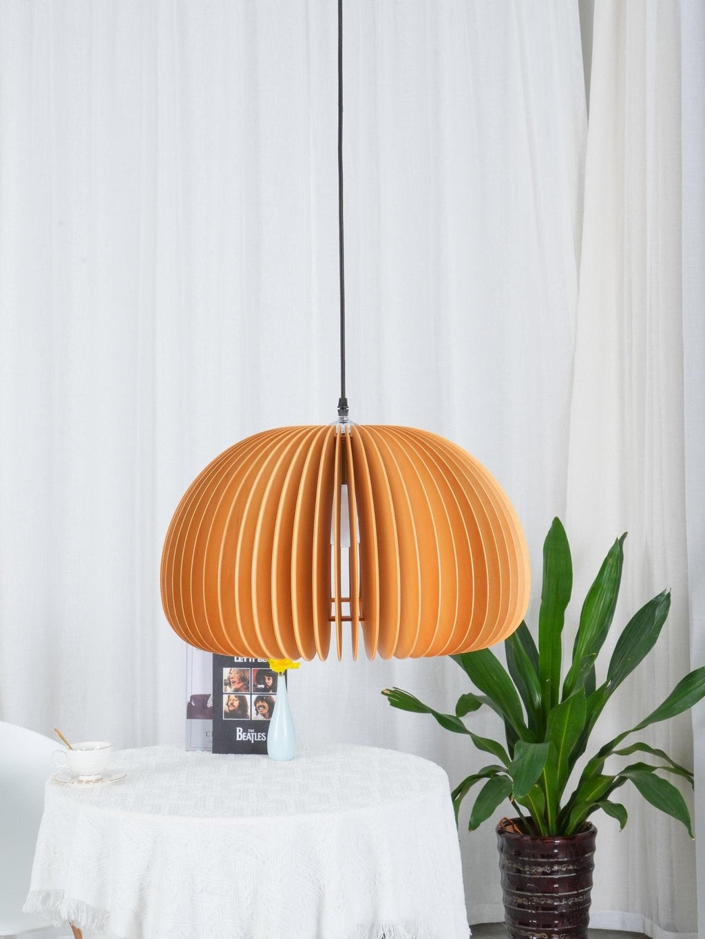 Clérachic slatted dome pendant in front of white curtains, with a table of books and a large leafy plant at the right.