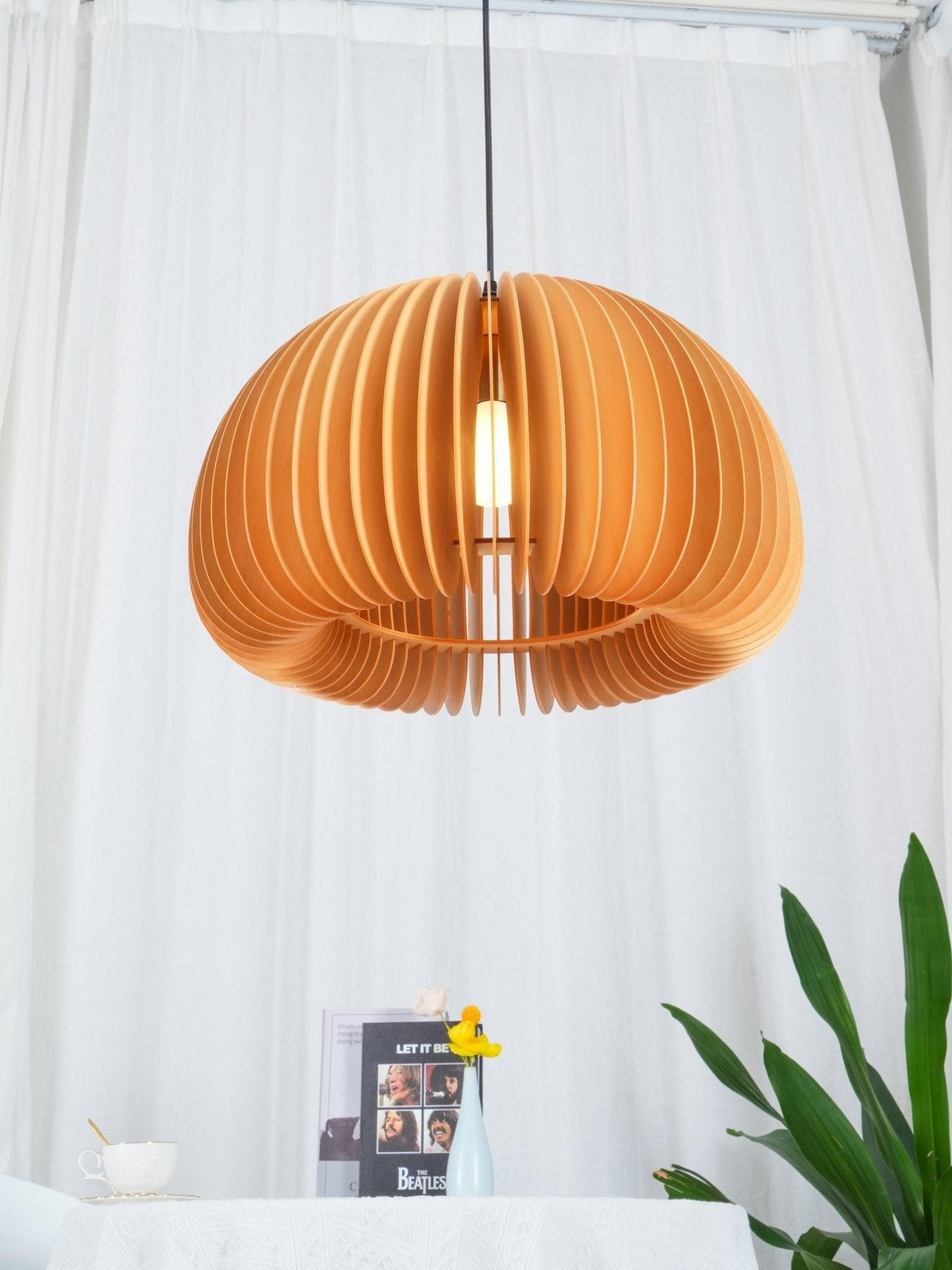 Clérachic wood pendant lit in front of billowing white curtains, with a plant, a small book and a cup on a table below.