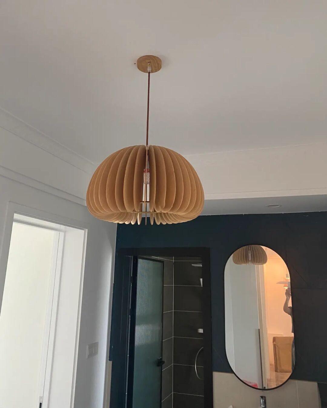 A Clérachic natural wood pendant with a wooden ceiling plate, hung in a white room with a tall mirror and a dark bathroom door.