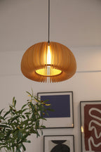 A Clérachic wood slat pendant lit against a pale wall, with the glow of the bulb showing through the open base and artwork below.