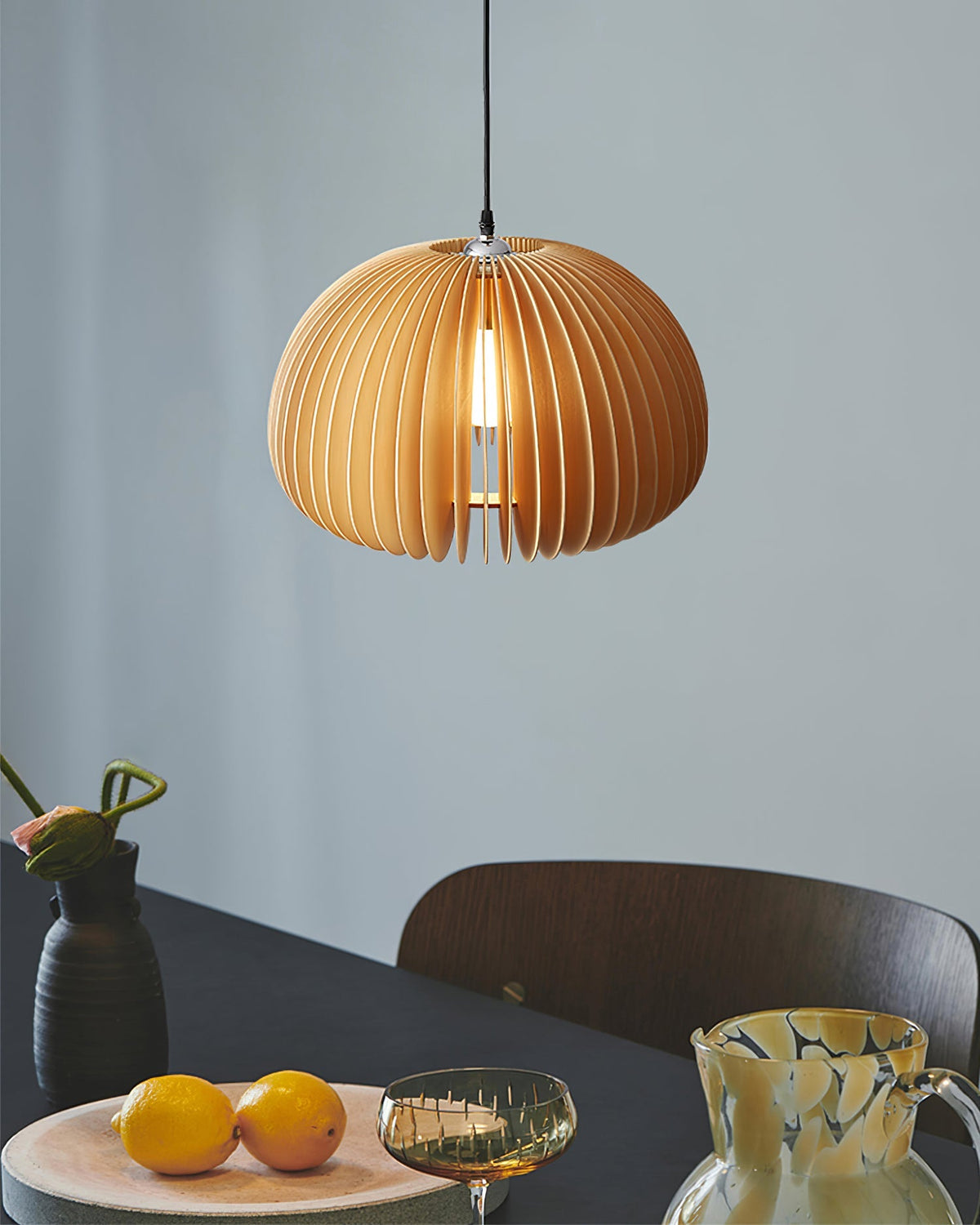 This Clérachic wooden pendant lit against a blue-grey wall, above a dark table with a bowl of oranges and a patterned jug.