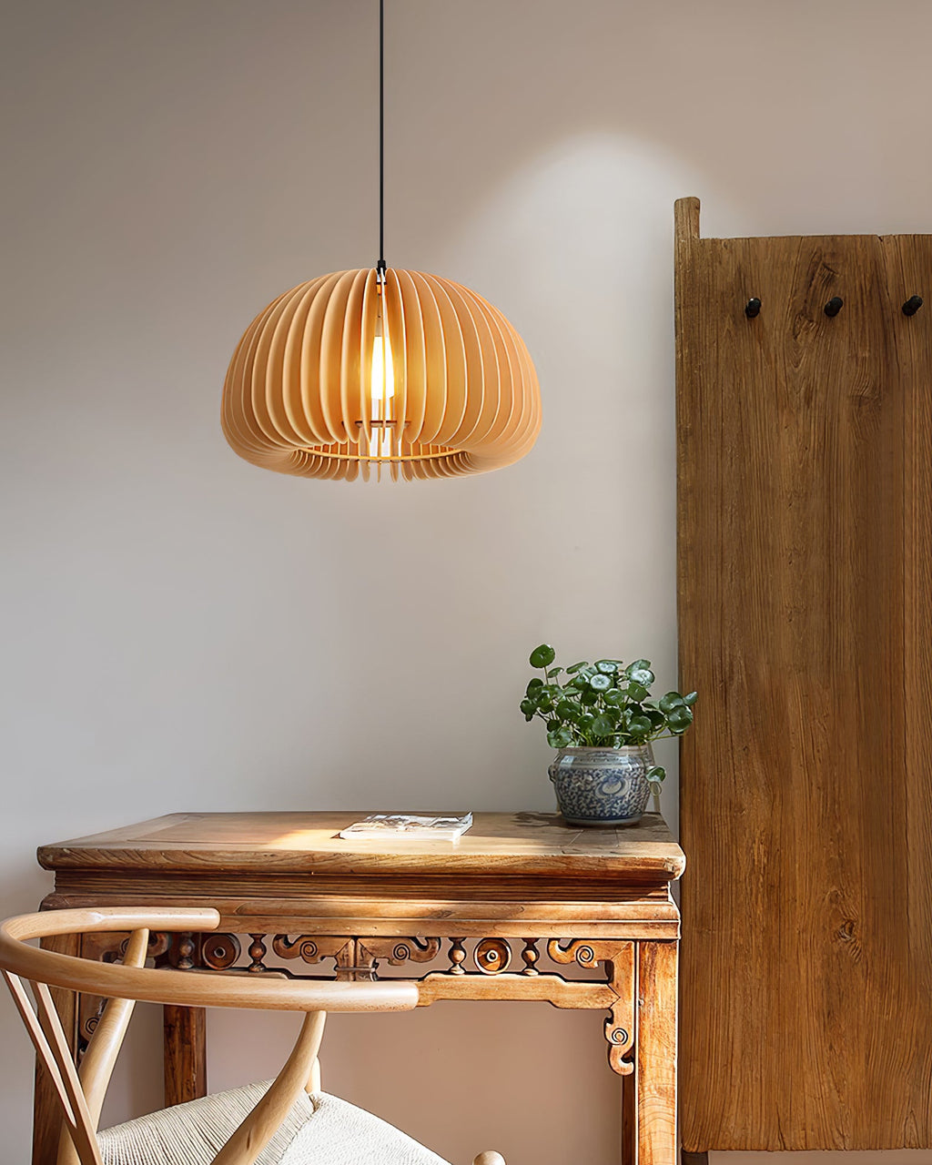 A Clérachic dome-shaped slatted pendant hung above a carved wooden console, with a small plant and a tall plank of wood at the side.