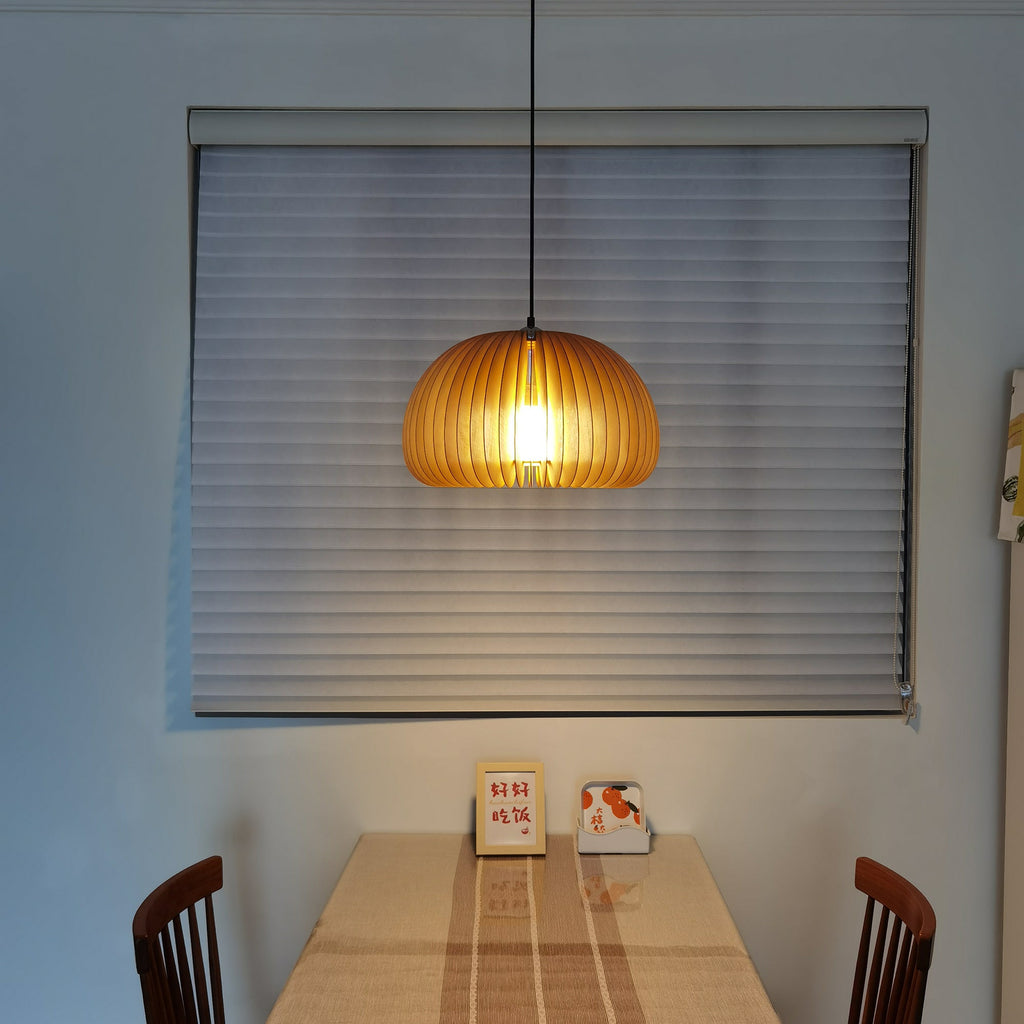 Clérachic wooden pendant lit in front of a grey window blind, above a table with a small framed card and wooden chairs.