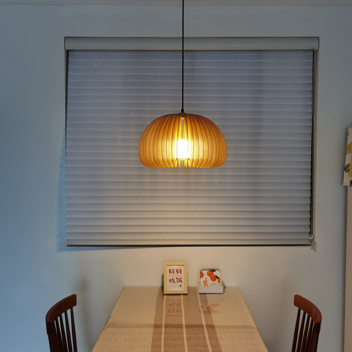 Clérachic wooden pendant lit in front of a grey window blind, above a table with a small framed card and wooden chairs.