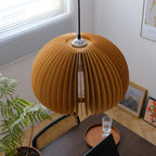 Clérachic slatted pendant seen from above, hanging over a dark table with a wooden tray, a glass and a green fern by a window.