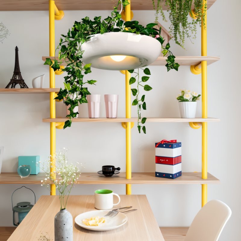 Clérachic white planter-style pendant with ivy hung before open yellow-pipe shelving, above a light wood table with a plate and mug.