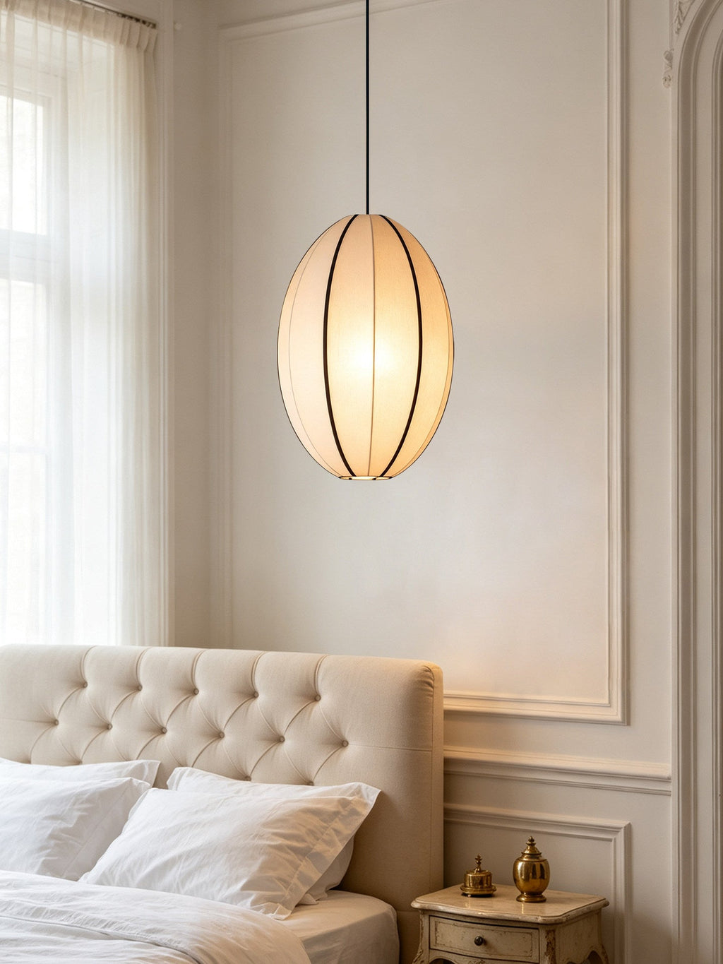 This Clérachic pendant's vertical oval shade hangs beside a tufted headboard in a pale bedroom.