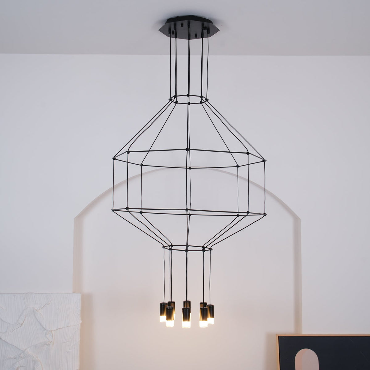 Lit black wireframe pendant on a round ceiling canopy, with a tiered cage and eight small lights below, against a white arched wall, from Clérachic.