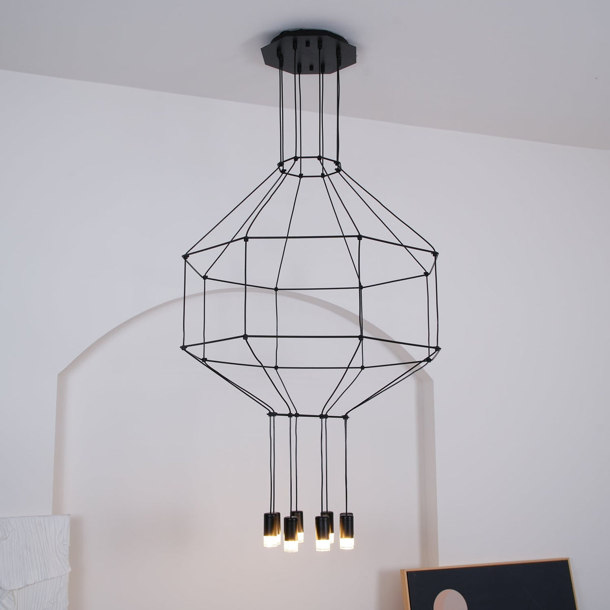 Clérachic wireframe pendant with a black canopy and tiered cable cage, its small lights glowing beneath, against a white wall.